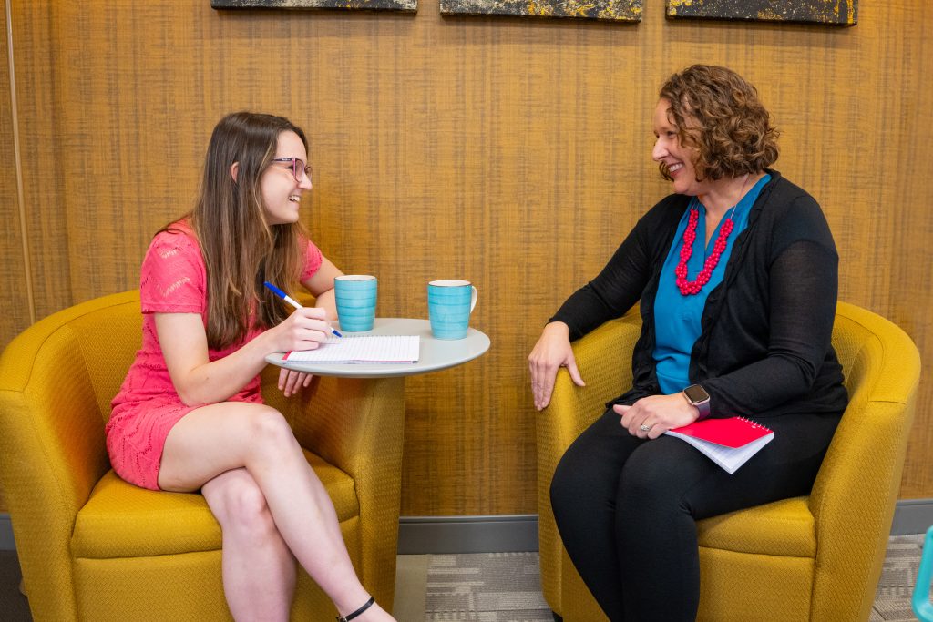 Two women are seated in yellow armchairs, engaged in a conversation. One is writing in a notebook on a small round table with blue mugs in front of them, likely discussing aspects of the complex sales process.