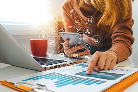 Woman in front of laptop pointing to data charts document while holding smart phone with marketing concepts icons around